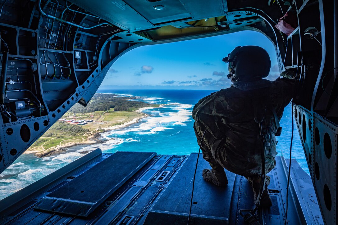 A solider kneels while looking out of the rear of a military helicopter as it flies over a beach on a sunny day.
