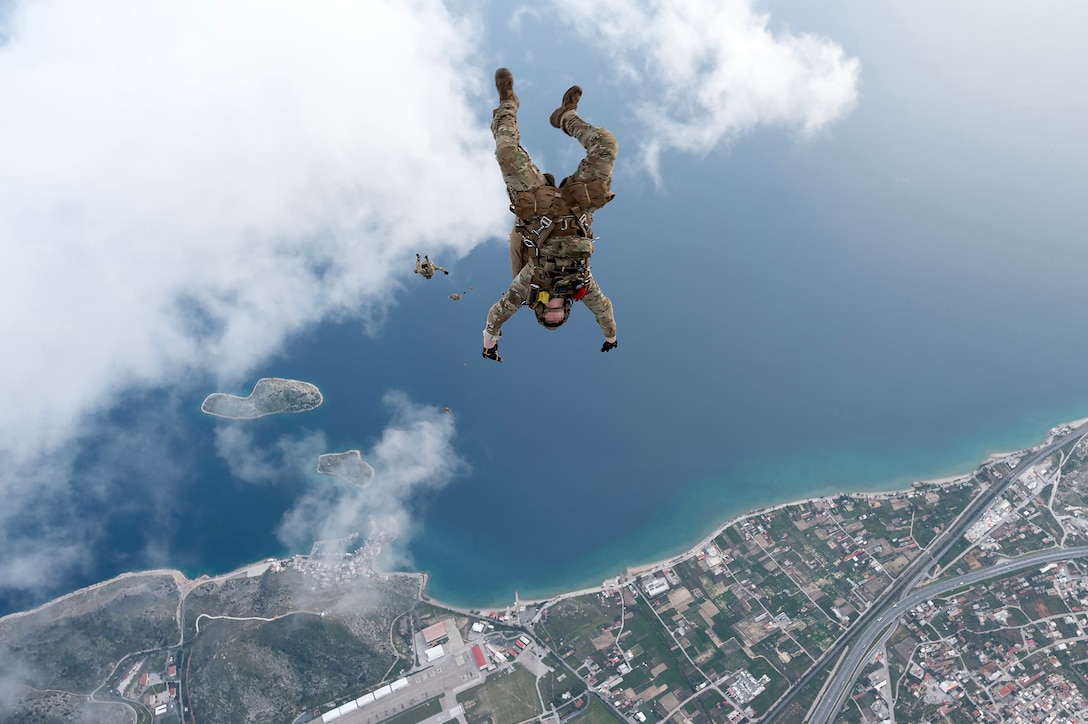 Service members jump headfirst from a military aircraft while in flight. Clouds and a city’s shoreline are beneath them.