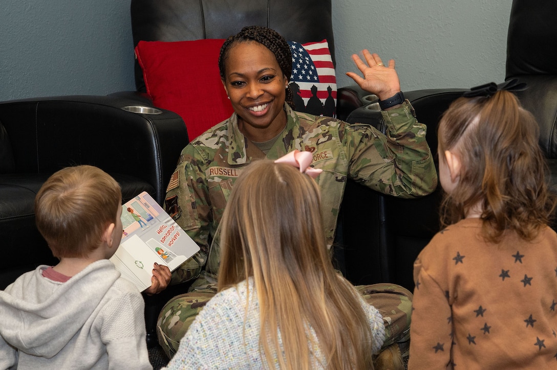 A service member holds a book in one hand and gestures with the other while reading to three seated children.