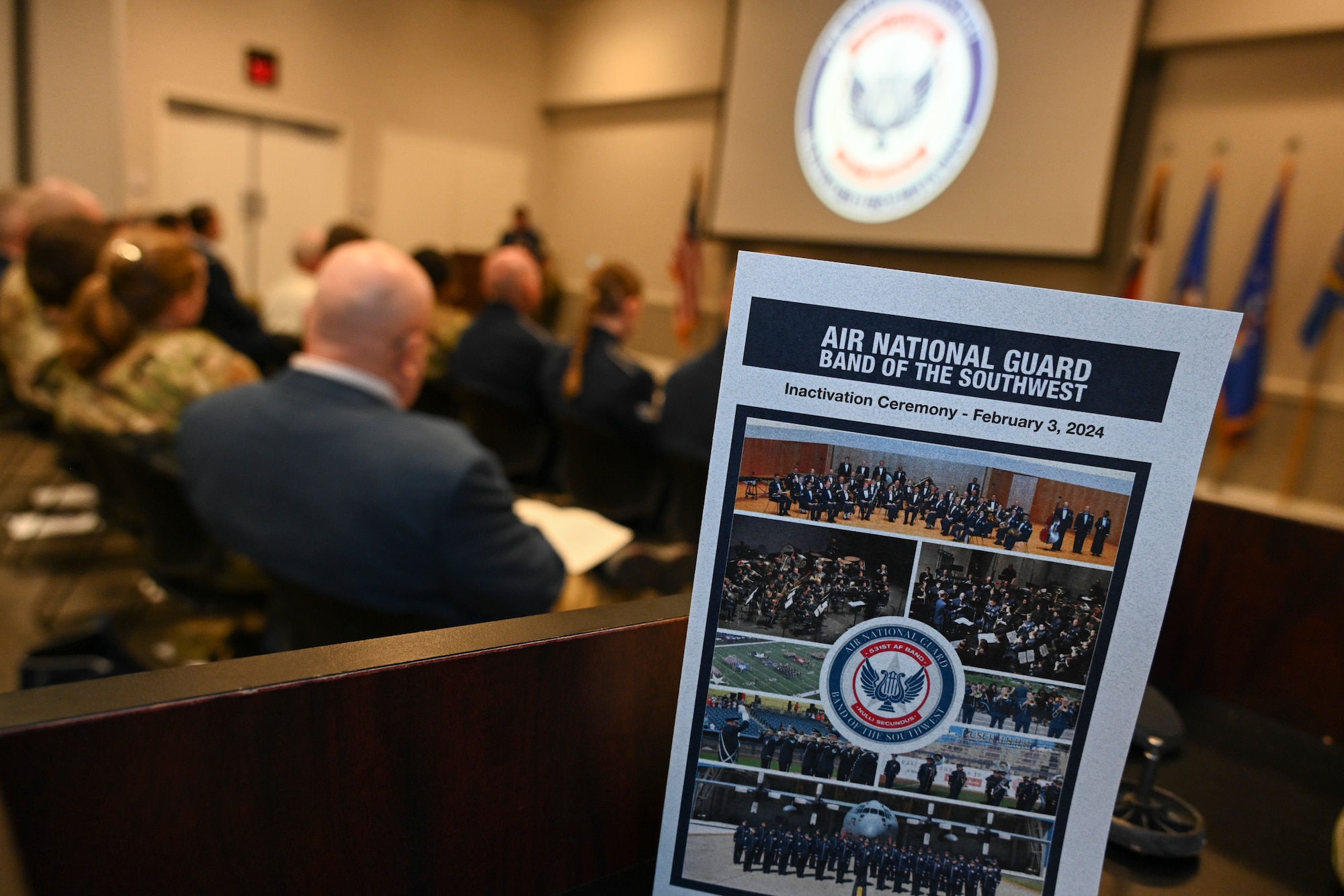 Foreground had a ceremony flyer, out of focus background has seated audience members listening to a speaker.