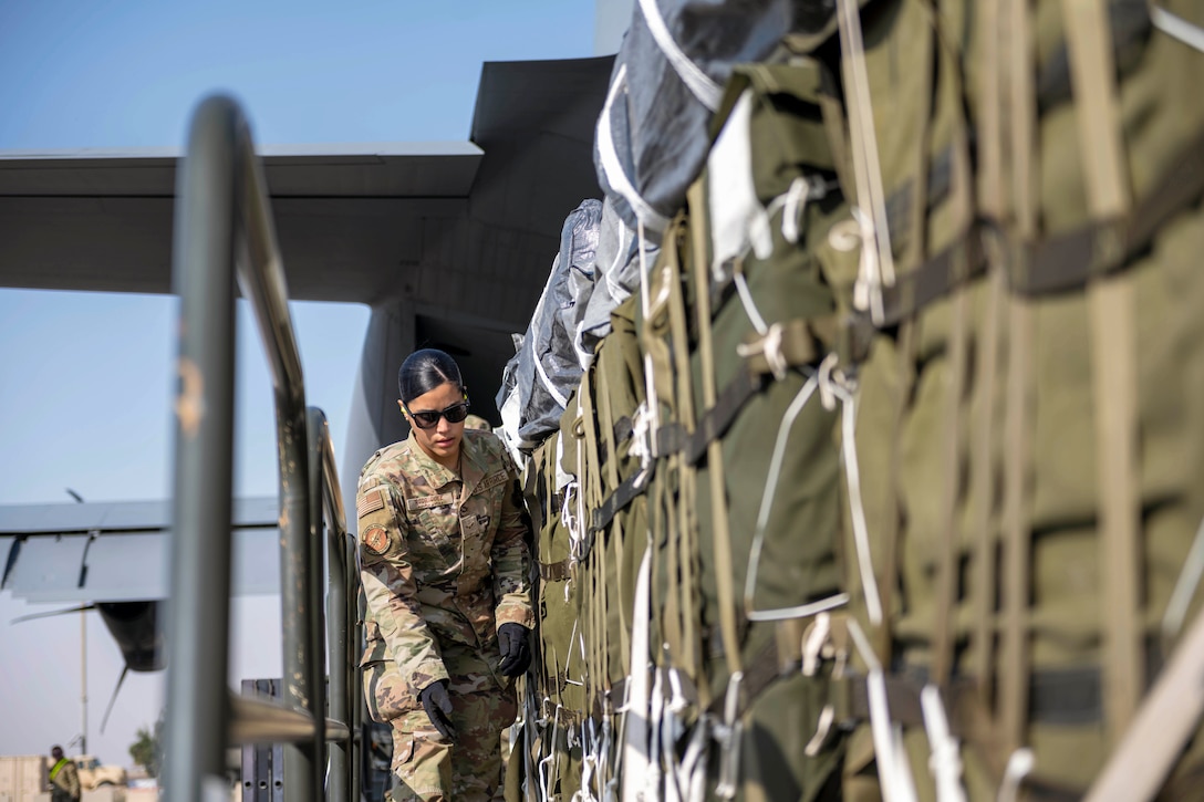 An airman walks next to bundles of a cargo as it enters an aircraft partially displayed in the background.