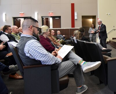 Photo is of a man standing and gesturing to a group of people in an auditorium