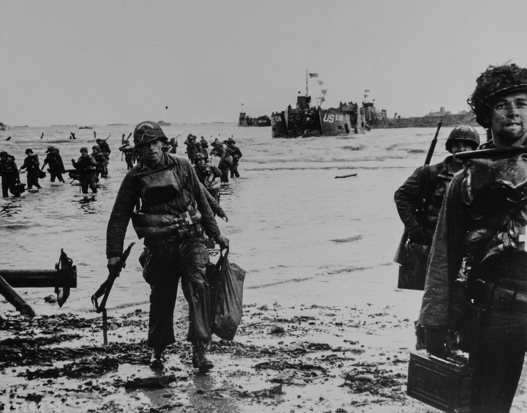Troops wade through surf and walk onto the shore with a landing craft in the background.