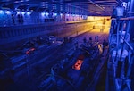 Service members stand around two small boats in a darkened well deck of a ship under purple lights.