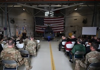 An audience sits in rows on the left and right of the screen, framing a grey helicopter displayed in the center.