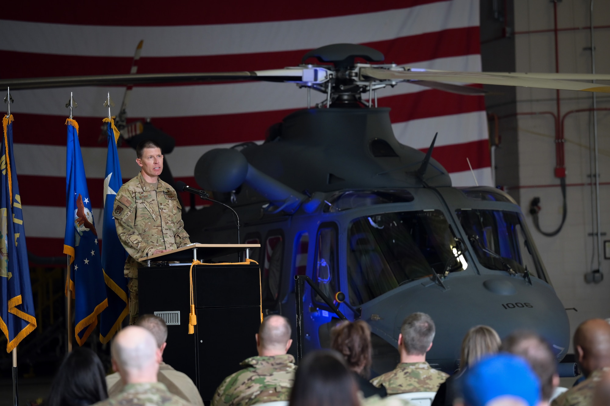 A man in military uniform stands behind a podium and speaks into a microphone as he addresses a seated crowd in an aircraft hangar.
