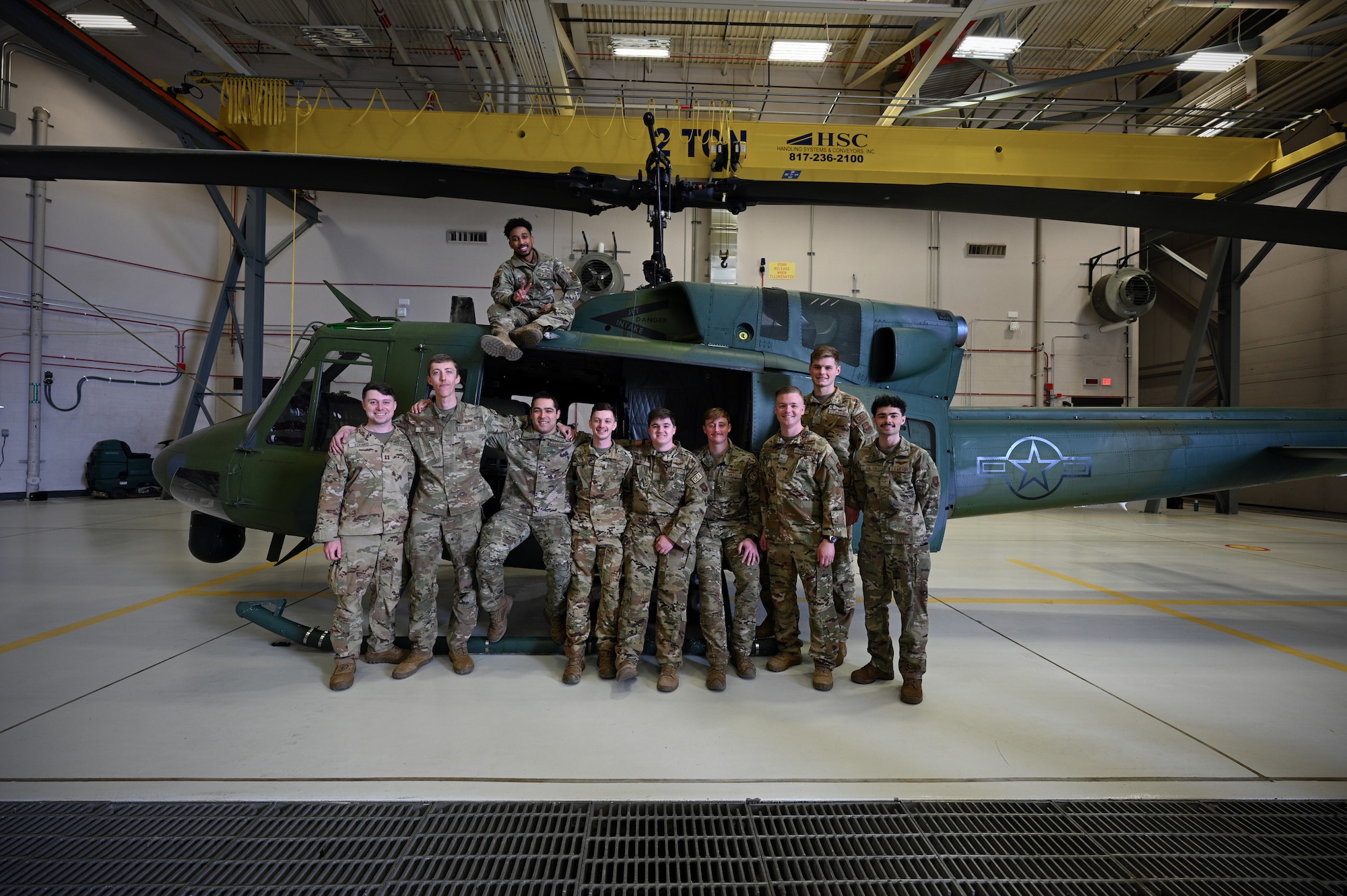 A group of ten uniformed military members stand next to a green helicopter in an aircraft hangar.
