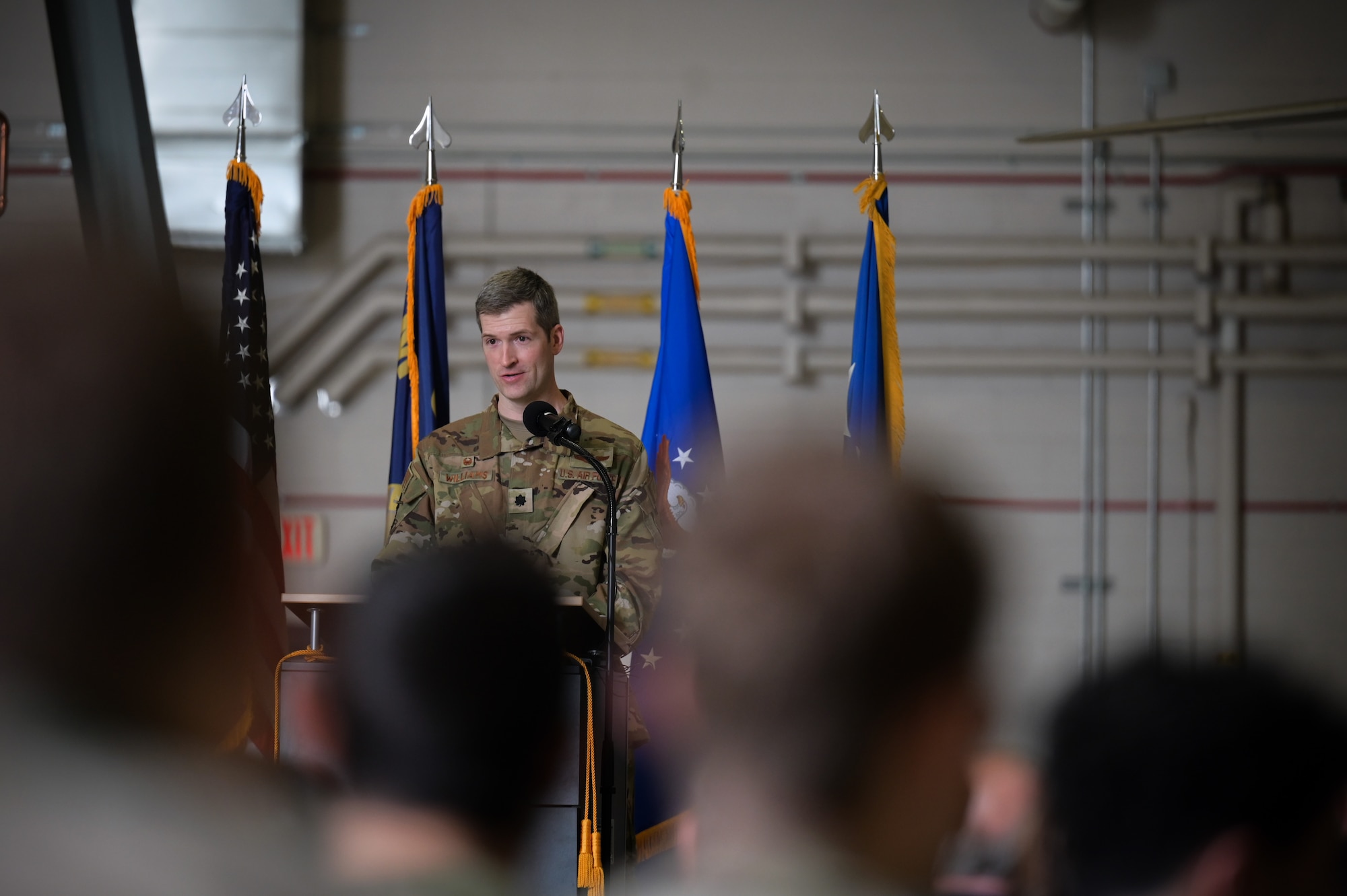 A man in military uniform stands in front of four blue flags with yellow tassles.