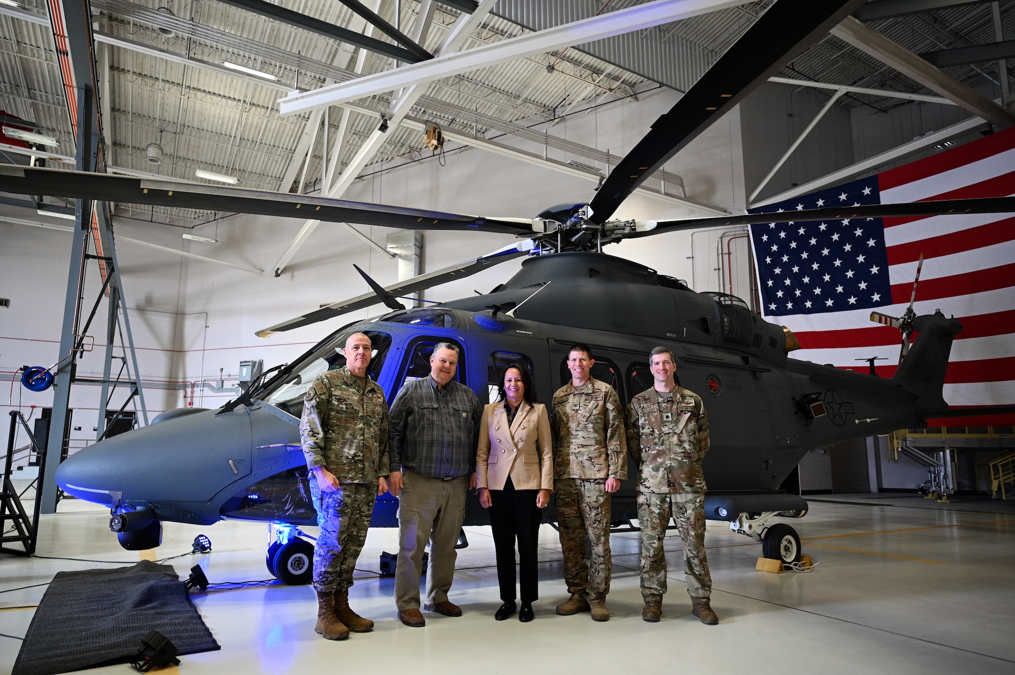 A group of five adults, three in military uniform and two in business clothes, stand beside each other and smile toward the camera. Behind them is a grey helicopter and large American flag.