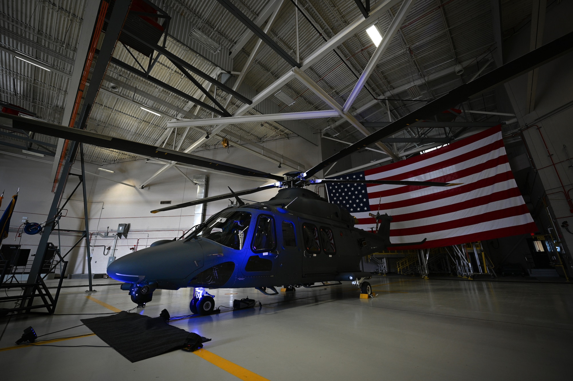 A grey helicopter is displayed in an aircraft hangar with a large American flag behind it.