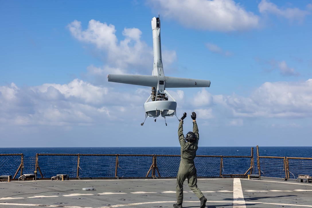 An uncrewed aerial vehicle launches from a ship while a service member stands on the deck with arms raised.