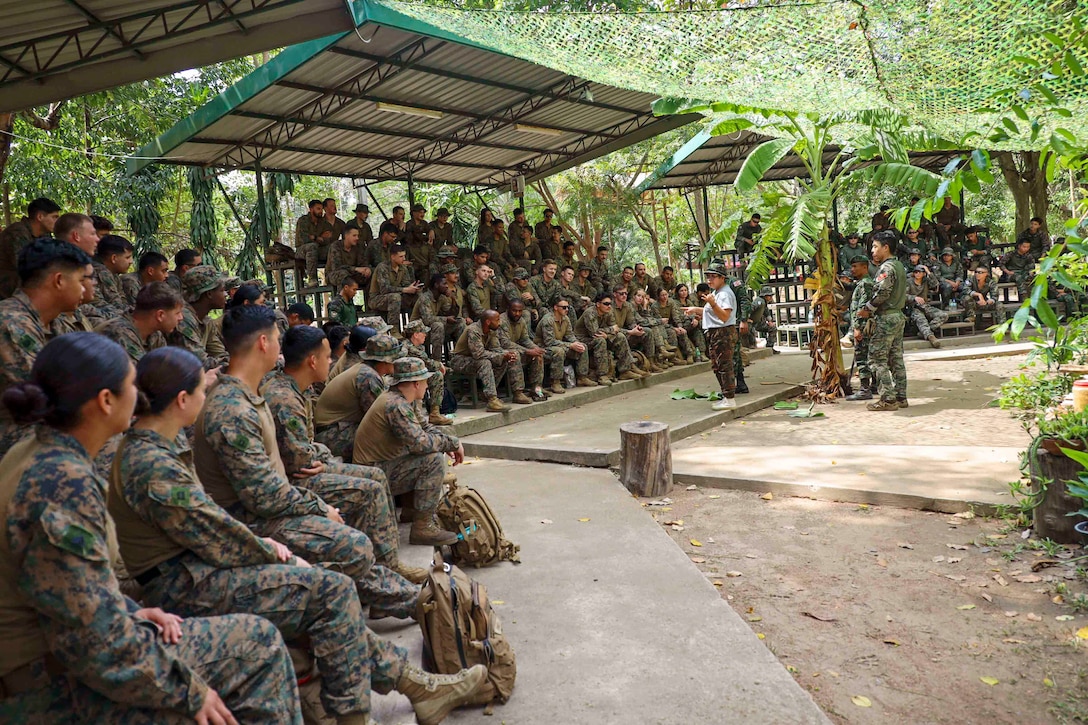 Groups of Marines sit on benches covered by metal roofs and listen to another Marine speaking in a jungle environment.