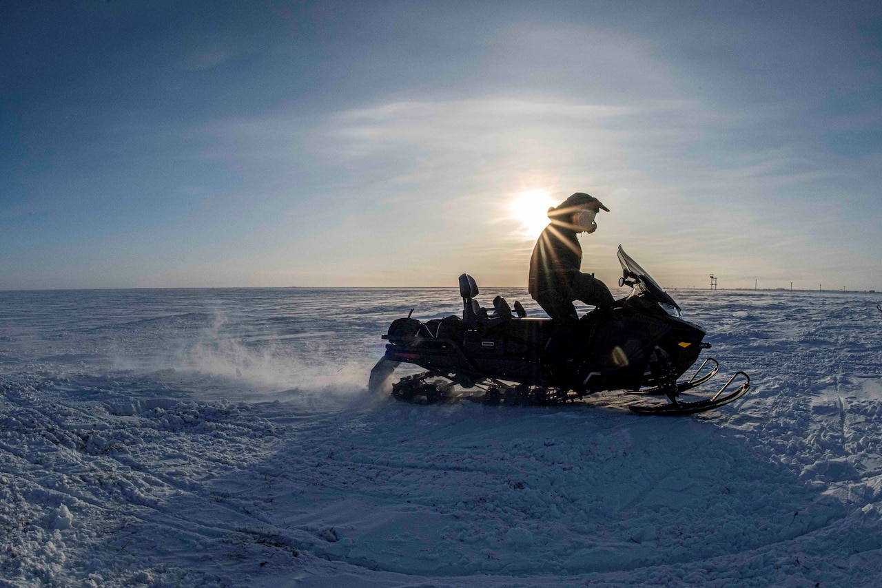 A service member operates a snowmobile on snow ground with a low sun in the background.