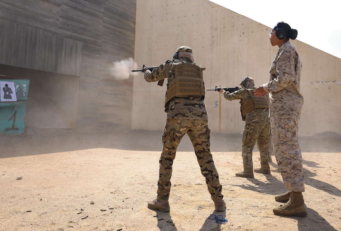 Two service members fire at targets in a desert area during marksmanship training as a third service member observes.