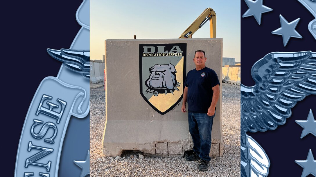 Man in a black t-shirt and blue jeans stands outside next to a concrete barrier taller than himself. on the barrier is a DLA logo with a dog's head. The photo was taken in a warm dry climate.