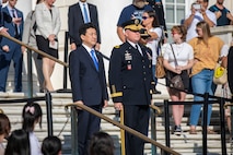 The Japanese defense minister is standing next to an Army General on some steps. They both have their hands at their sides. There are people dressed in various types of clothes all around them watching the ceremony.