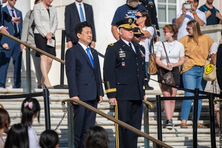 The Japanese defense minister is standing next to an Army General on some steps. They both have their hands at their sides. There are people dressed in various types of clothes all around them watching the ceremony.