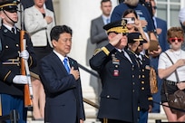 The Japanese defense minister is wearing a dark suit and bright blue tie, and he has his hand over his heart while the general next to him and others are saluting. The audience in the background is also holding their hands over their hearts.
