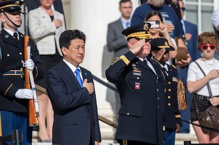 The Japanese defense minister is wearing a dark suit and bright blue tie, and he has his hand over his heart while the general next to him and others are saluting. The audience in the background is also holding their hands over their hearts.