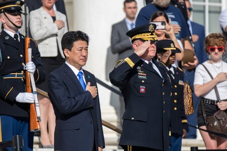 The Japanese defense minister is wearing a dark suit and bright blue tie, and he has his hand over his heart while the general next to him and others are saluting. The audience in the background is also holding their hands over their hearts.