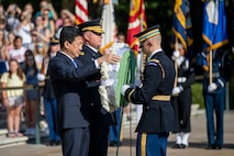 The Japanese Minister of Defense is dressed in a dark suit, and his is placing a wreath on a stand while an Army general stands next to him. An Army soldier is assisting him with the wreath.
