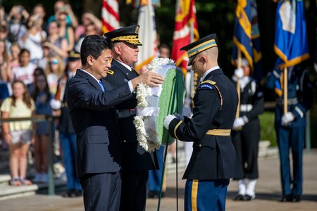 The Japanese Minister of Defense is dressed in a dark suit, and his is placing a wreath on a stand while an Army general stands next to him. An Army soldier is assisting him with the wreath.