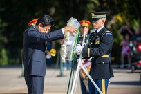 The Japanese minister of defense is placing a wreath onto a stand while an Army soldier assists him.