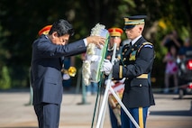 The Japanese minister of defense is placing a wreath onto a stand while an Army soldier assists him.