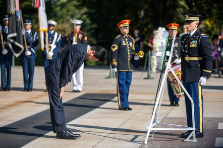 The Japanese minister of defense is dressed in a dark suit, and his is bowing in front of a wreath as a US Army Soldier stands at attention with hands at his sides near the wreath. In the distance is an Army band member holding a trumpet under one arm, and service member carrying service flags.
