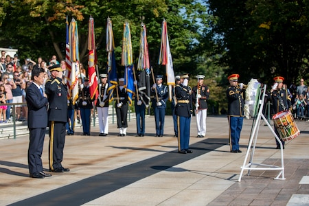 The Japanese minister of defense has his hand over his heart and an Army general officer is saluting a wreath, while a member of the Army band is seen in the distance with a trumpet to play Taps. A drummer is standing next to him saluting. There is also a color guard holding the US flag and the flags of each branch of the military in the background.