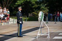 A soldier has a sword in his hand so the tip is pointing up near his shoulder. He is standing in front of a white wreath, and there are people standing on white marble steps in the background.