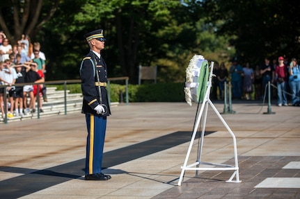 A soldier has a sword in his hand so the tip is pointing up near his shoulder. He is standing in front of a white wreath, and there are people standing on white marble steps in the background.