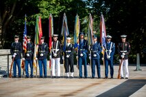 Nine service members from each branch of the military are standing in a row holding the US flag and the flags of each branch of the military.