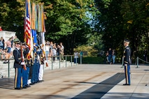 A color guard of service members is standing with the US Flag and the flags of the branches of the military in front of the Tomb of the Unknown Soldier. Another Soldier is standing at a distance in front of them, holding a sword in his hand that is pointed to the ground.