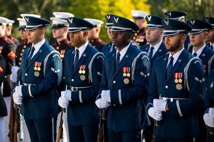 Members of the US Air Force Honor Guard are standing at rest with their ceremonial rifles in front of them with the butt of the rifle on the ground. They are dressed in Air Force blue uniforms