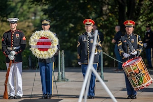 Four military members are standing. The Marine on the left has a rifle in front of him, resting the end on the ground. The Soldier next to him is holding the wreath on a stand. The other two service members pictured are in the Army band. One is carrying a trumpet under his arm and the other is carrying a large drum in front of him.