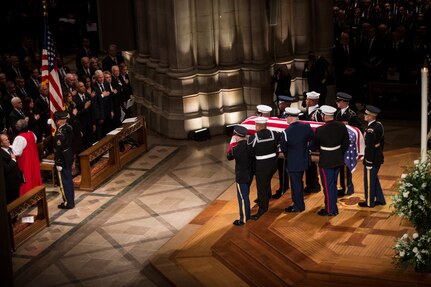 Military members are carrying a casket off the wood platform at Washington National Cathedral. They are heading toward the main aisle, following a soldier who is carrying the US flag.