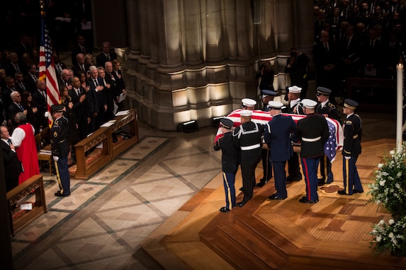 Military members are carrying a casket off the wood platform at Washington National Cathedral. They are heading toward the main aisle, following a soldier who is carrying the US flag.