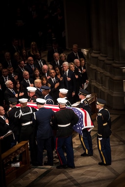 Service members are carrying a flag-draped casket down the aisle at Washington National Cathedral while former presidents and their spouses and other audience members look on with their hands over their hearts.