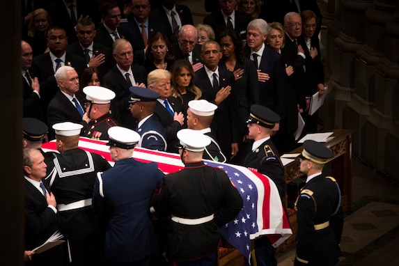 Service members are carrying a flag-draped casket down the aisle of Washington National Cathedral. Former presidents and their spouses, and other audience members are holding their hands over the hearts while watching the procession.