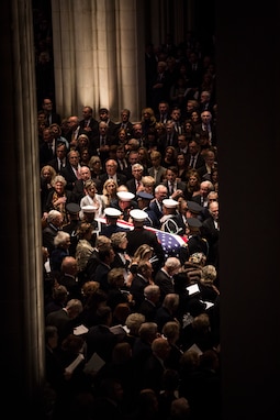 Service members from each branch of the military dressed in their ceremonial uniforms are carrying a flag-draped casket down the middle aisle of the Washington National Cathedral as the congregation is standing and watching the procession.