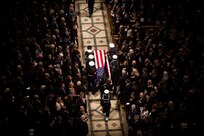 Overhead shot of military members are carrying a flag-draped casket down the center aisle of Washington National Cathedral. On either side of them are full rows of people dressed in dark colors.