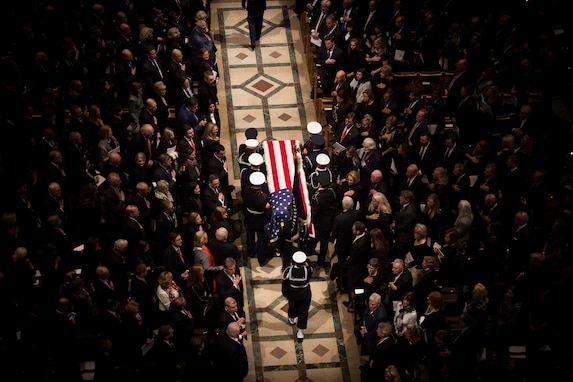 Overhead shot of military members are carrying a flag-draped casket down the center aisle of Washington National Cathedral. On either side of them are full rows of people dressed in dark colors.