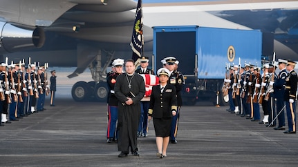 Service members are carrying a flag-draped casket away from Air Force One, while other service members are flanking them on the flightline.