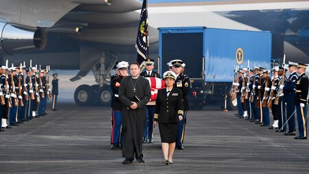 Service members are carrying a flag-draped casket away from Air Force One, while other service members are flanking them on the flightline.
