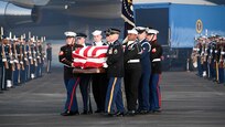 Service members are carrying a flag-draped casket away from Air Force One, while other service members are flanking them on the flightline.