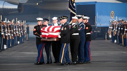 Service members are carrying a flag-draped casket away from Air Force One, while other service members are flanking them on the flightline.