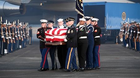 Service members are carrying a flag-draped casket away from Air Force One, while other service members are flanking them on the flightline.