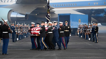 Service members are carrying a flag-draped casket away from Air Force One, while other service members are flanking them on the flightline.