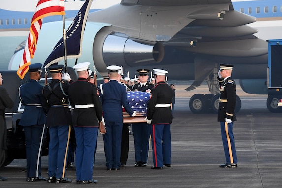 Members of the military are putting a flag-draped casket into a hearse. In the distance is Air Force One. One service members is saluting and others are holding the US flag and the Flag of the US President.